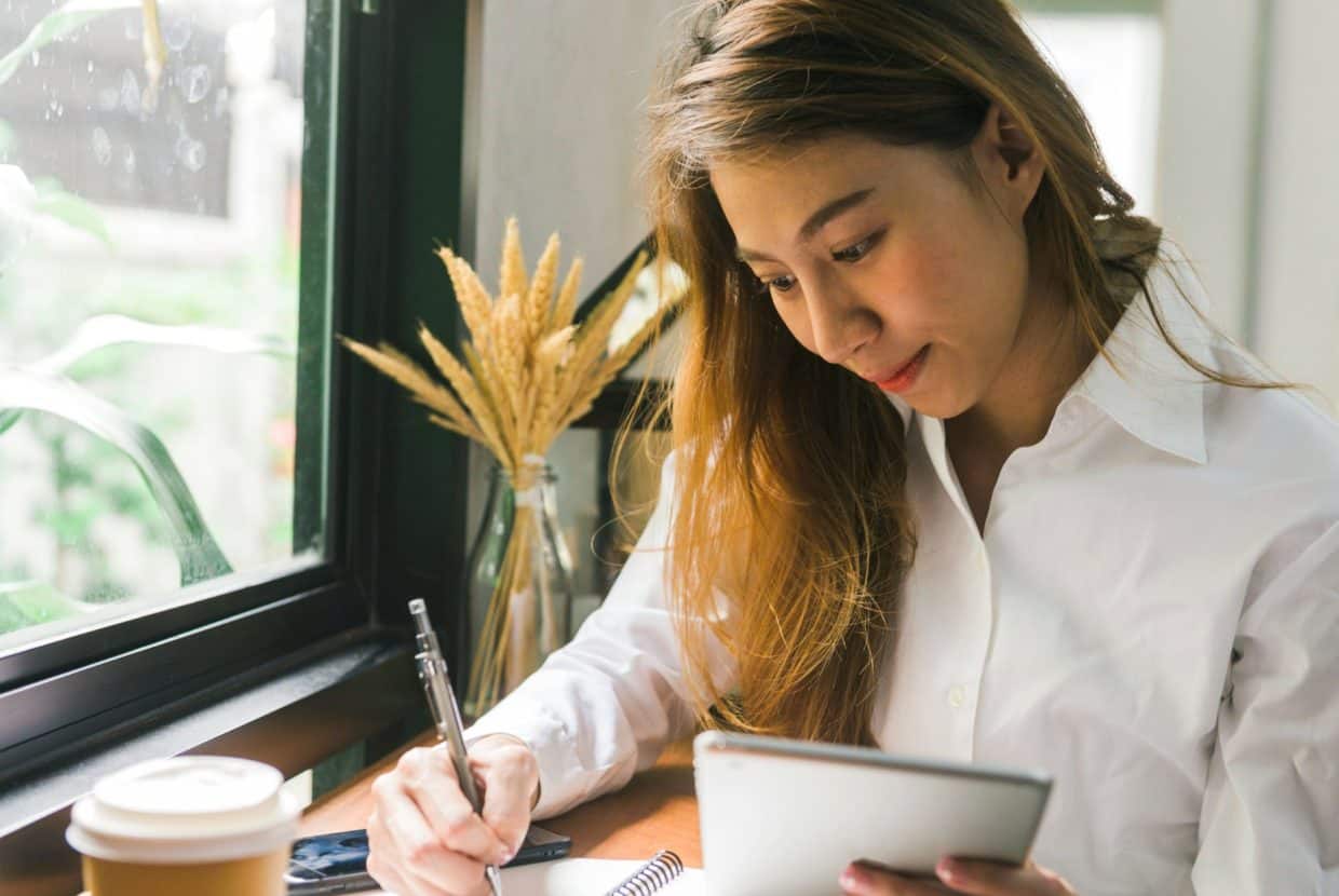 Young business woman writing in a notebook while holding a tablet by a window with coffee nearby.