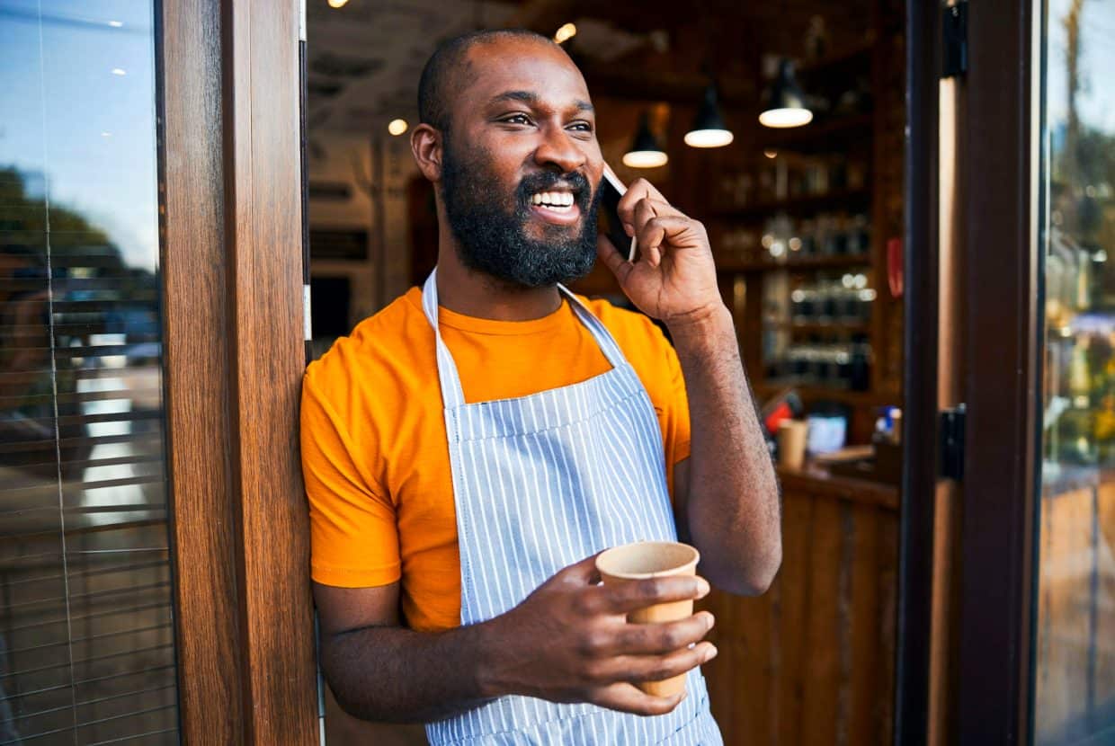 Smiling business owner in apron talking on the phone and holding a coffee outside his café.