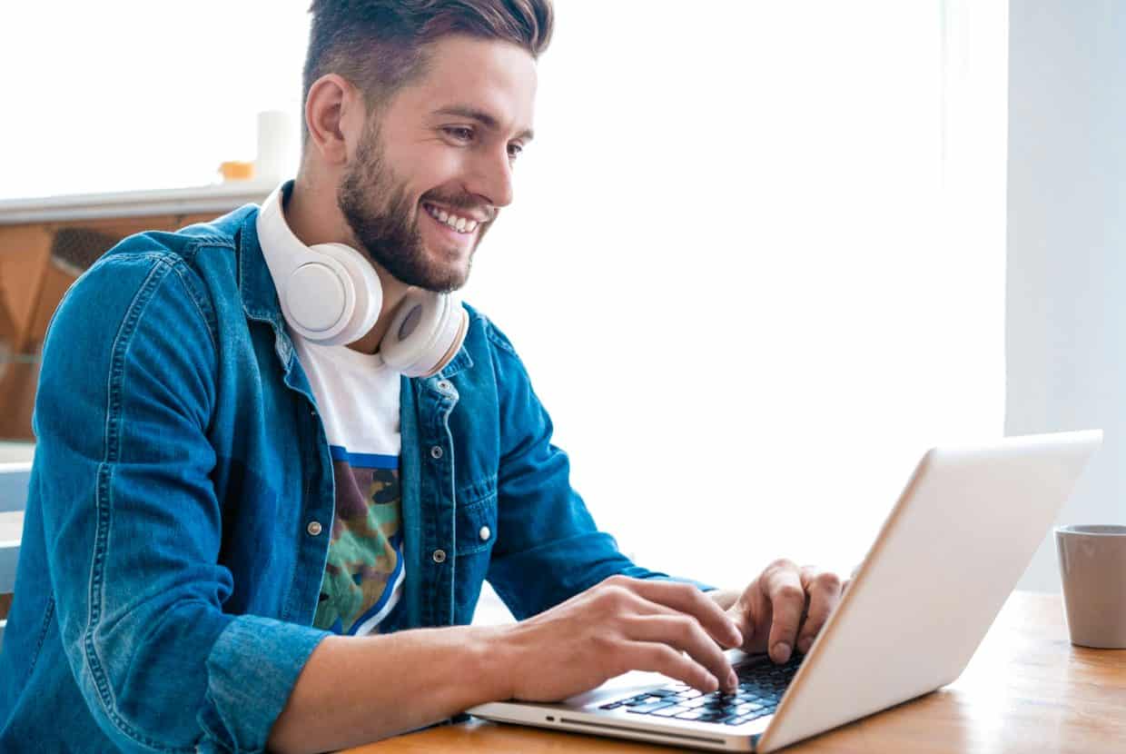 Man smiling while working on a laptop, with headphones resting around his neck.