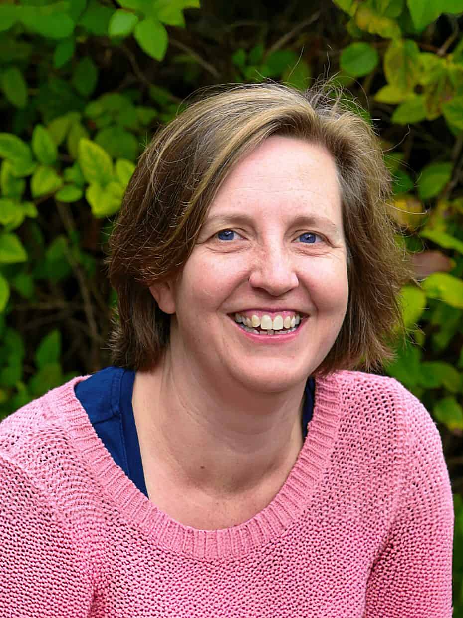 Smiling woman wearing a pink jumper sitting outdoors in front of green leafy bushes.