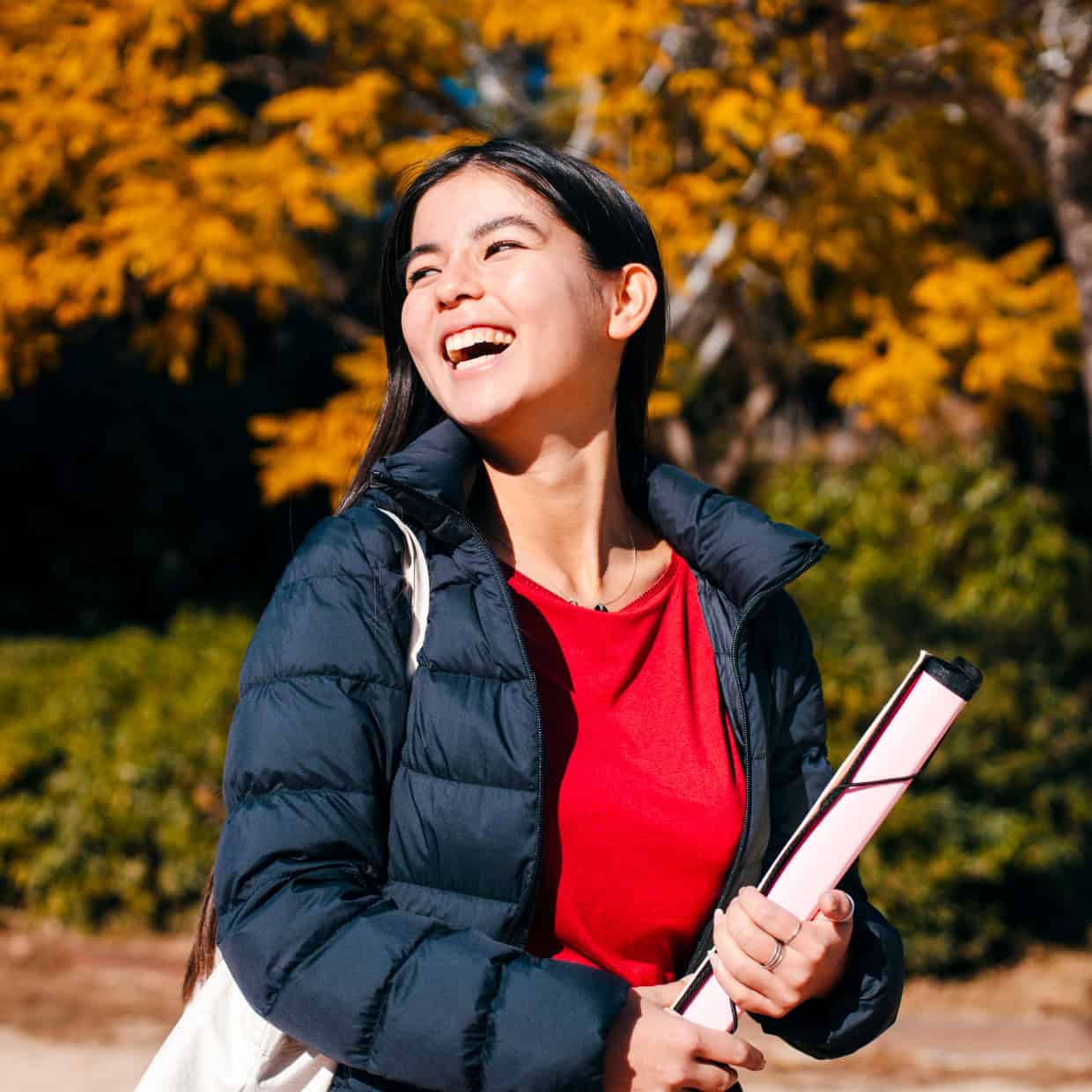 A smiling young woman in a red top and puffer jacket holding a rolled-up poster outdoors in autumn.