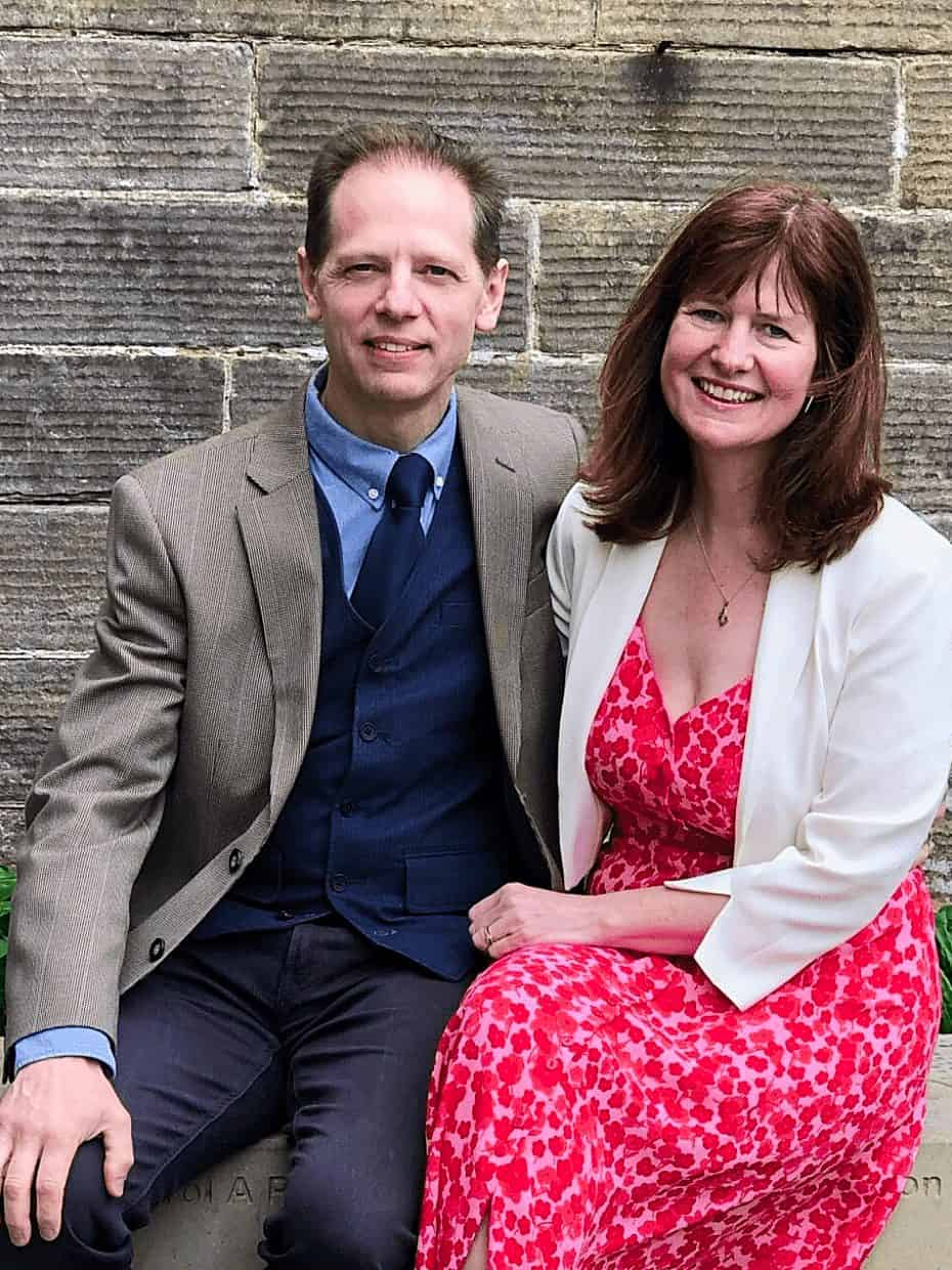 Smiling couple sitting together on a stone bench in front of a brick wall, dressed smartly in a suit and a pink floral dress.