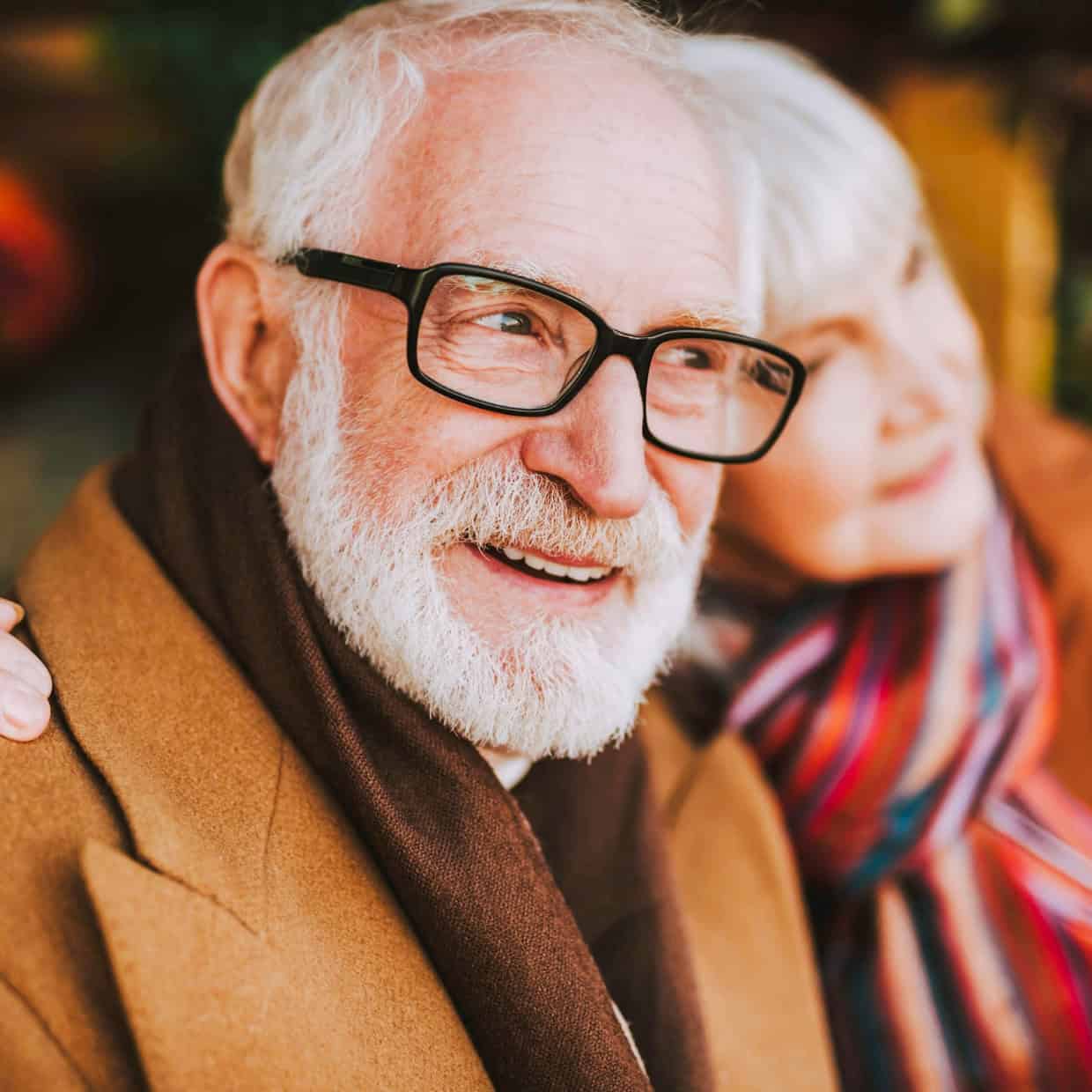 Smiling man wearing glasses and a coat, with an elderly woman beside him leaning in affectionately.