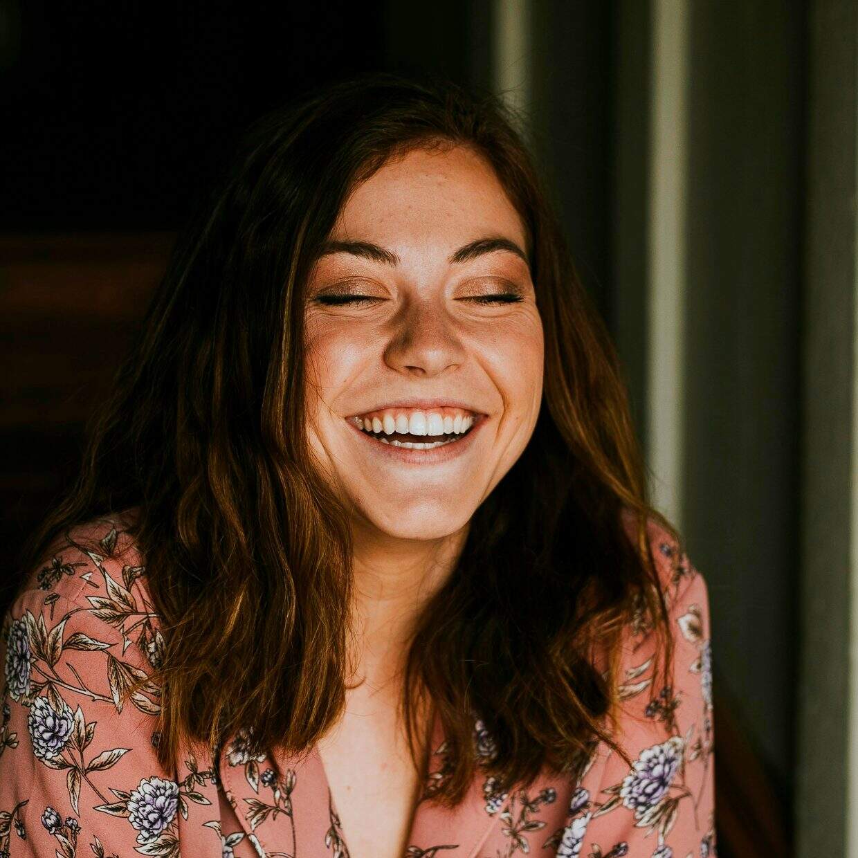 Young woman in floral top smiling with eyes closed, sitting indoors.