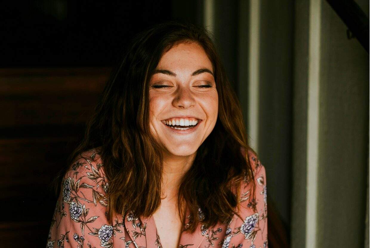 Young woman in floral top smiling with eyes closed, sitting indoors.