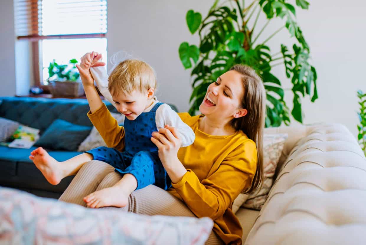 Mother and young child playing happily on a sofa at home with plants in the background.