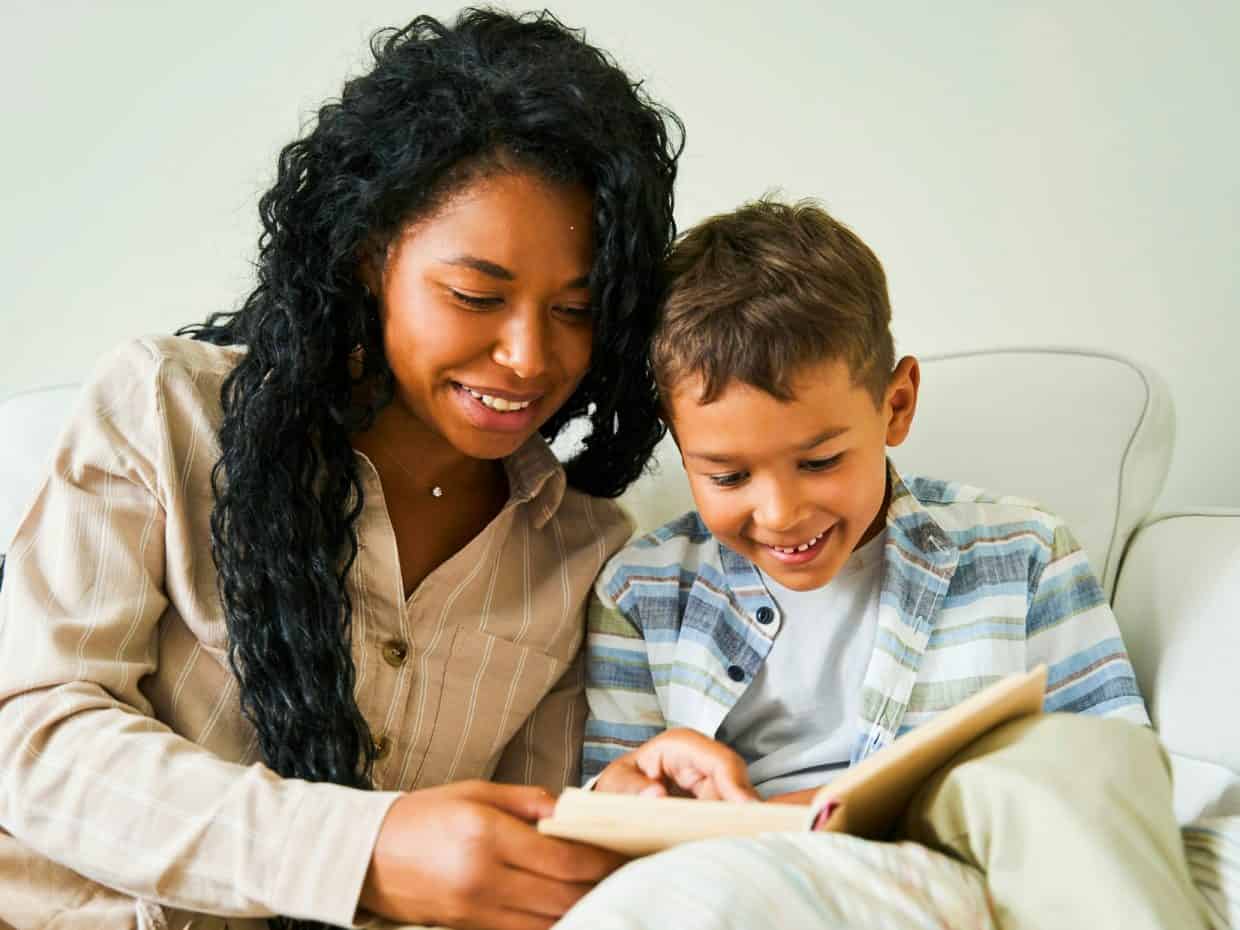 Smiling woman reading a book with a young boy on a white sofa.